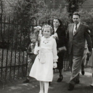 dziewczynka w białej sukience i wianku na głowie idzie ulicą, za nią idą mały chłopiec, kobieta i mężczyzna w marynarce