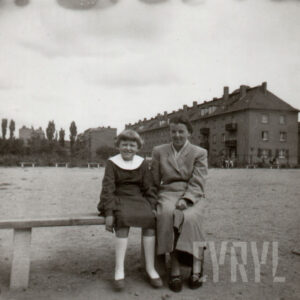 na ławce siedzi mała dziewczynka w ciemnej sukience z białym kołnierzykiem i w białych podkolanówkach, obok niej siedzi kobieta w płaszczu, w głębi dom ze spadzistym dachem