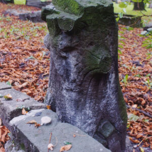 ścięty, nieokorowany pień drzewa ustawiony u wezgłowia ziemnej mogiły, pień z wystającymi ściętymi sękami o zaznaczonych słojach, z przodu pnia płaskorzeźba przedstawiająca motyw liści i owoców dębu, pień drzewa pokryty zielonym nalotem