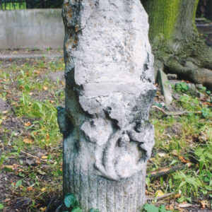 ścięty, nieokorowany pień drzewa z wystającymi ściętymi sękami o zaznaczonych słojach, z przodu pnia płaskorzeźba przedstawiająca motyw liści i owoców dębu, nad dekoracją ścięte ukośnie miejsce, na której pierwotnie była umieszczona tablica inskrypcyjna