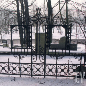 mogiła ziemna ogrodzona kutym, metalowym ogrodzeniem osadzonym na niskiej granitowej podmurówce, w części zagłówkowej pośrodku kuty żeliwny krzyż o trójlistnie zakończonych ramionach z rozetką pośrodku, po bokach dwa prostokątne obramienia tablic inskrypcyjnych (brak tablic) , w głębi kamienne nagrobki i drzewa; sceneria zimowa