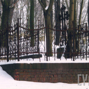 mogiła ziemna ogrodzona ozdobną żeliwną kutą kratą, osadzoną na kamiennym cokole wspartym na ceglanym podmurowaniu, pośrodku, z prawej metalowy, ażurowy krzyż łaciński z trójlistnie zakończonymi ramionami, w głębi drzewa, sceneria zimowa