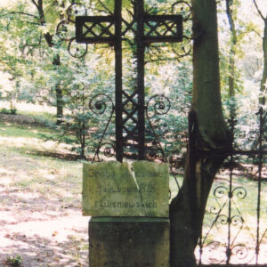 metalowy, ażurowy krzyż łaciński z trójlistnie zakończonymi ramionami, wypełniony ukośnymi krzyżykami, umieszczony na cokole z piaskowca z marmurową pękniętą tablicą w kształcie otwartej księgi z wyrytym napisem, z prawej fragment kutej ozdobnej kraty i pień drzewa