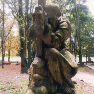 rzeźba klęczącego, starszego mężczyzny w długiej szacie, z kapeluszem na plecach obejmującego krzyż stojący na postumencie, widok od strony twarzy mężczyzny