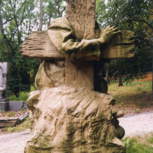 rzeźba krzyża od tyłu, za nim zarys postaci klęczącego, starszego mężczyzny (głowa niewidoczna) w długiej szacie, obejmującego jedną ręką krzyż