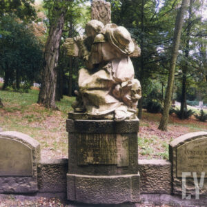 nagrobek w formie tryptyku, pośrodku rzeźba starszego mężczyzny w długiej szacie, z kapeluszem na plecach obejmującego krzyż stojący na postumencie, po bokach pionowe płyty z inskrypcjami zamknięte łukiem