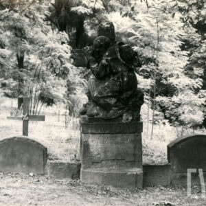 nagrobek w formie tryptyku, pośrodku rzeźba starszego mężczyzny w długiej szacie, z kapeluszem na plecach obejmującego krzyż stojący na postumencie, po bokach pionowe płyty zamknięte łukiem