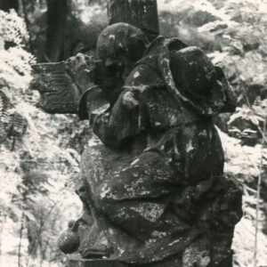 rzeźba starszego, klęczącego mężczyzny w długiej szacie, z kapeluszem na plecach obejmującego krzyż stojący na postumencie