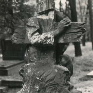 rzeźba krzyża od tyłu, za nim zarys postaci klęczącego, starszego mężczyzny w długiej szacie, obejmującego jedną ręką krzyż