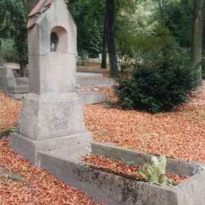 mogiła ziemna obramowana wąskimi ściankami porośniętymi mchem, w partii zagłówkowej murowana kapliczka stylizowana na przydrożną, zamknięta trójkątnym szczytem nakrytym dwuspadowym daszkiem pokrytym dachówką, na jego szczycie metalowy krzyż, w górnej części kapliczki płytka nisza