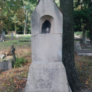 murowana kapliczka stylizowana na przydrożną, zamknięta trójkątnym szczytem nakrytym dwuspadowym daszkiem, na jego szczycie metalowy krzyż, w górnej części kapliczki płytka nisza, poniżej zatarta inskrypcja, z prawej pień drzewa, w głębi murowane nagrobki