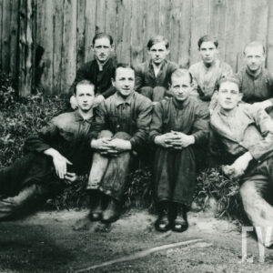 grupa młodych mężczyzn w ubraniach roboczych, siedzą w dwóch rzędach na ziemi, w tle drewniane ogrodzenie