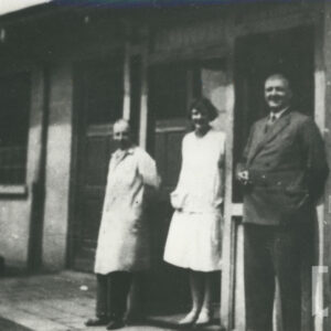 trójka osób, jeden mężczyzna w garniturze, kobieta w sukience, mężczyzna w stroju roboczym, stojący przed murowanym budynkiem z oknami i drzwiami wejściowymi