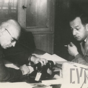 dwóch starszych mężczyzn pochylających się nad stołem z dokumentami i telefonem 