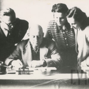 starszy mężczyzna siedzący przy biurku, wokół niego stoją młodzi ludzie - mężczyzna i dwie kobiety