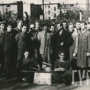 kilkunastu mężczyzn, część w strojach roboczych, część w płaszczach, pozujących do fotografii; dwóch z nich trzyma silnik z kartką informującą o wyprodukowaniu 1000 egzemplarzy urządzeń