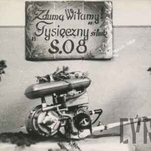 urządzenie mechaniczne leżące na podeście; u góry wisi kartka z napisem: Z dumą Witamy Tysięczny silnik S.08