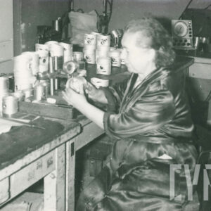 starsza kobieta w ubraniu roboczym, siedząca przy biurku i trzymająca w dłoniach metalowy przedmiot