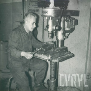 starszy mężczyzna w ubraniu roboczym, siedzący przy stanowisku z wiertarką