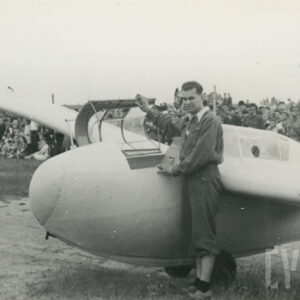 mężczyzna w stroju lotnika przy samolocie leżącym na trawiastym lądowisku, w głębi widzowie