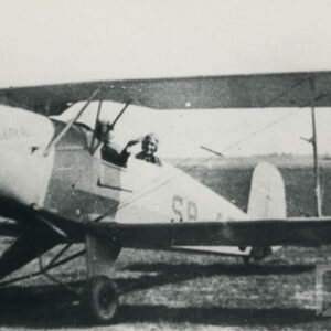 mężczyzna w stroju pilota, salutujący z kokpitu samolotu stojącego na trawiastym lądowisku 