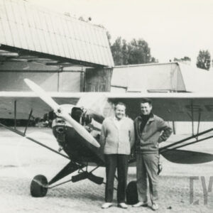 dwóch mężczyzn stojących przy samolocie na tle hangarów samolotowych