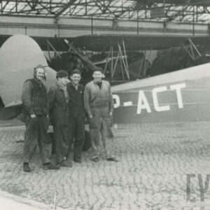 czterech mężczyzn stojących przy samolocie, trzech z nich nosi ubrania robocze, jeden strój pilota, w głębi inne samoloty stojące wewnątrz hangaru