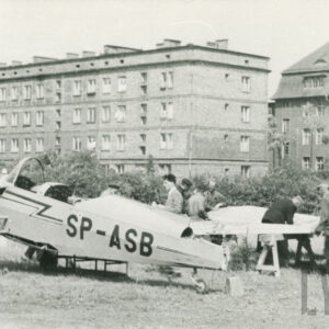 grupa mężczyzn przyglądająca się rozłożonemu na części samolotowi ustawionemu na trawiastym terenie, w tle bloki mieszkalne i kamienica z cegieł