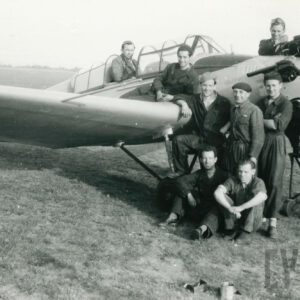 grupa mężczyzn w strojach roboczych pozująca do zdjęcia przy samolocie stojącym na trawiastym lądowisku