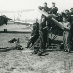 grupa mężczyzn w strojach roboczych pozująca do zdjęcia przy samolocie stojącym na trawiastym lądowisku