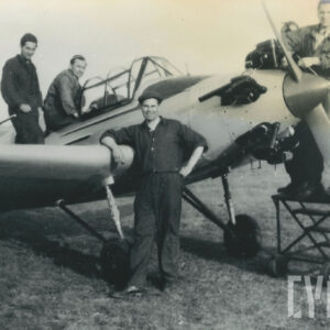 grupa mężczyzn w strojach roboczych pozująca do zdjęcia przy samolocie stojącym na trawiastym lądowisku