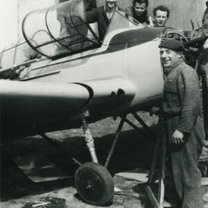 grupa mężczyzn w strojach roboczych pozująca do zdjęcia przy samolocie stojącym na trawiastym lądowisku