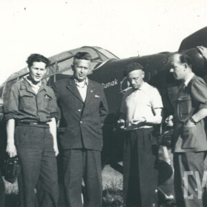 grupa mężczyzn w strojach roboczych pozująca do zdjęcia przy samolocie stojącym na trawiastym lądowisku