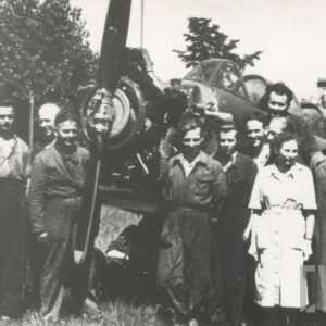 grupa osób w strojach roboczych pozująca do fotografii przy samolocie, w głębi drzewa i budynek mieszkalny