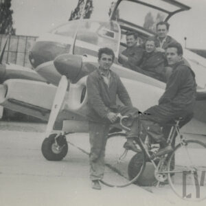 grupa mężczyzn pozująca do fotografii przy samolocie, trzech z nich siedzi w kokpicie, jeden stoi oparty o samolot, a jeden siedzi na rowerze i opiera się o skrzydło, w tle hangary