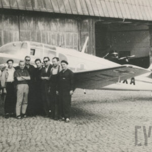 grupa mężczyzn opierających się o samolot i pozujących do fotografii, w tle hangar