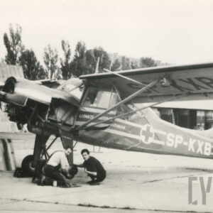 dwóch mężczyzn kucających przy kole podwozia samolotu, w tle hangar