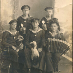 pięciu mężczyzn ubranych w wojskowe stroje i czapki marynarskie, pozujący na sztucznym tle w fotograficznym atelier; dwóch z nich stoi i trzyma w rękach kufle z piwem, trzech siedzi, jeden trzyma akordeon, jeden szmacianą laleczką, a ostatni z nich, siedzący pośrodku trzyma między nogami wypchanego ptaka
