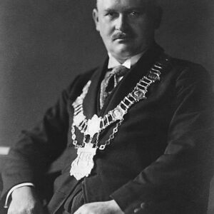 popiersie siedzącego mężczyzny w średnim wieku w białej koszuli, pod krawatem i ciemnej marynarce, na połach której spoczywa bogato zdobiony, okazały łańcuch