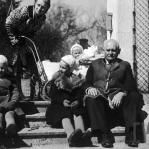 na schodach prowadzących do budynku siedzą starszy pan w grubej kufajce i dwoje małych dzieci, za nimi pochyla się kobieta, obok niej stoi wózek, a w nim dziecko w jasnej czapeczce