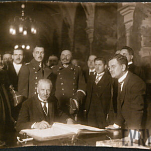 na pierwszym planie nad otwartą księgą leżącą na stole siedzi mężczyzna, wokół niego stoją inni mężczyźni, w tym wojskowi w galowych płaszczach, w głębi zarys zabytkowego pomieszczenia z kolumnami