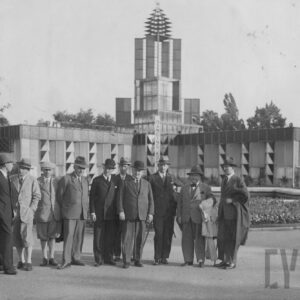 grupa mężczyzn w garniturach i kapeluszach oraz jedna kobieta stoją na tle modernistycznego budynku dwuskrzydłowego z wieżą