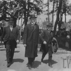 trzej mężczyźni w garniturach i płaszczach, dwóch melonikach, jeden w kapeluszu idą drogą, w tle szpaler ludzi i drzewa