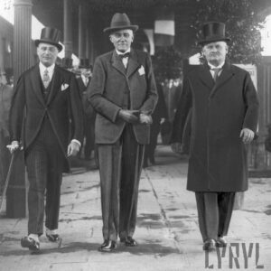 trzej mężczyźni, dwóch w cylindrach, jeden w kapeluszu, odświętnie ubrani, idą zadaszonym i podpartym żeliwnymi kolumnami peronem dworca kolejowego 