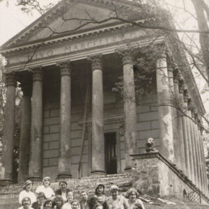 grupa młodych kobiet siedzi na trawniku przed wysoką, zwieńczoną tympanonem budowlą otoczoną kolumnadą