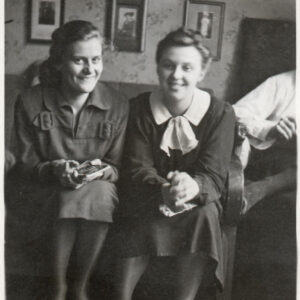 dwie uśmiechnięte, młode kobiety siedzą na kanapie w pokoju, którego ściany oklejone są wzorzystą tapetą, za ich plecami na ścianie wiszą zdjęcia oprawione w ramki