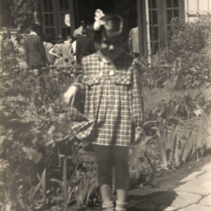 mała dziewczynka z kokardą we włosach i sukience w kratkę stoi na tle zielonych rabat, w głębi oszklone, wieloskrzydłowe drzwi