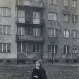 mała dziewczynka w czapce i płaszczyku stoi na trawniku przed czteropiętrowym blokiem mieszkalnym z balkonami