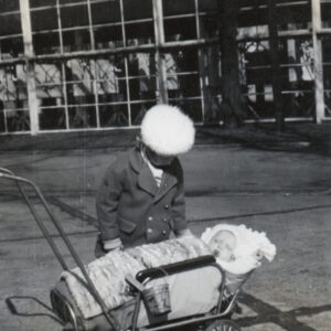 mała dziewczynka w płaszczyku i czapce pochyla się nad wózkiem z dzieckiem owiniętym w kocyk, w głębi fragment przeszklonego pawilonu