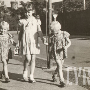 kilkuletnia dziewczynka w sukience z falbankami prowadzi za ręce ulicą dwie mniejsze dziewczynki w identycznych, wzorzystych sukienkach, w głębi zieleń i szczyty wież gmachów publicznych